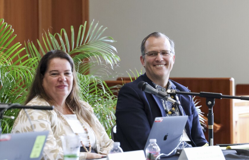 A woman and a man smile at a camera from behind the table they are seated at