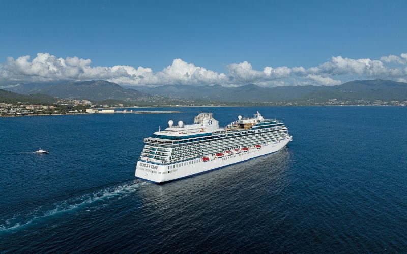 A large cruise ship sails on a calm blue ocean under a bright, clear sky. In the background, a coastal city lies at the base of green mountains, with scattered clouds adding texture to the sky.