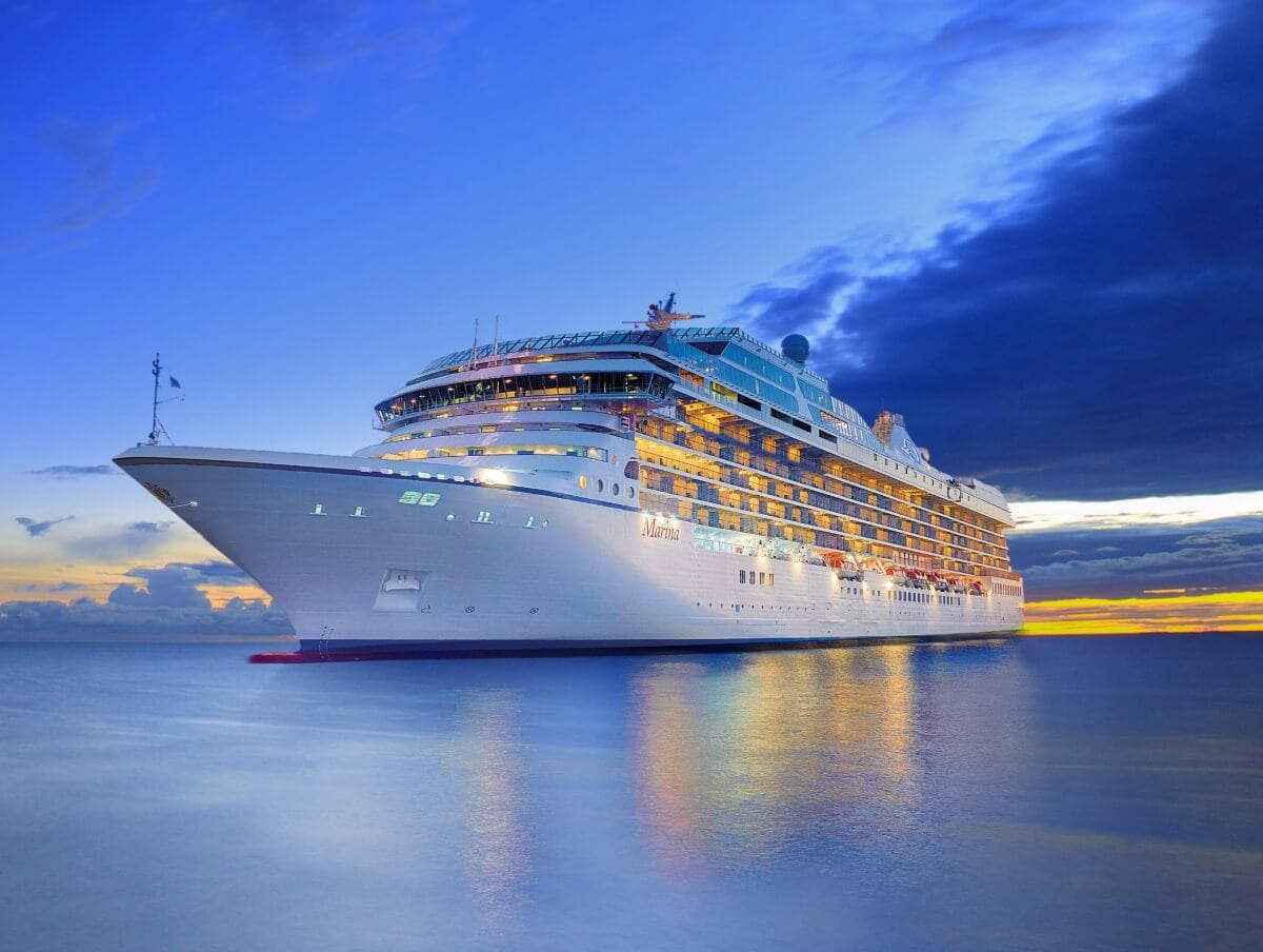 A large Oceania Cruises ship illuminated at dusk with a vibrant sunset in the background over calm sea waters.